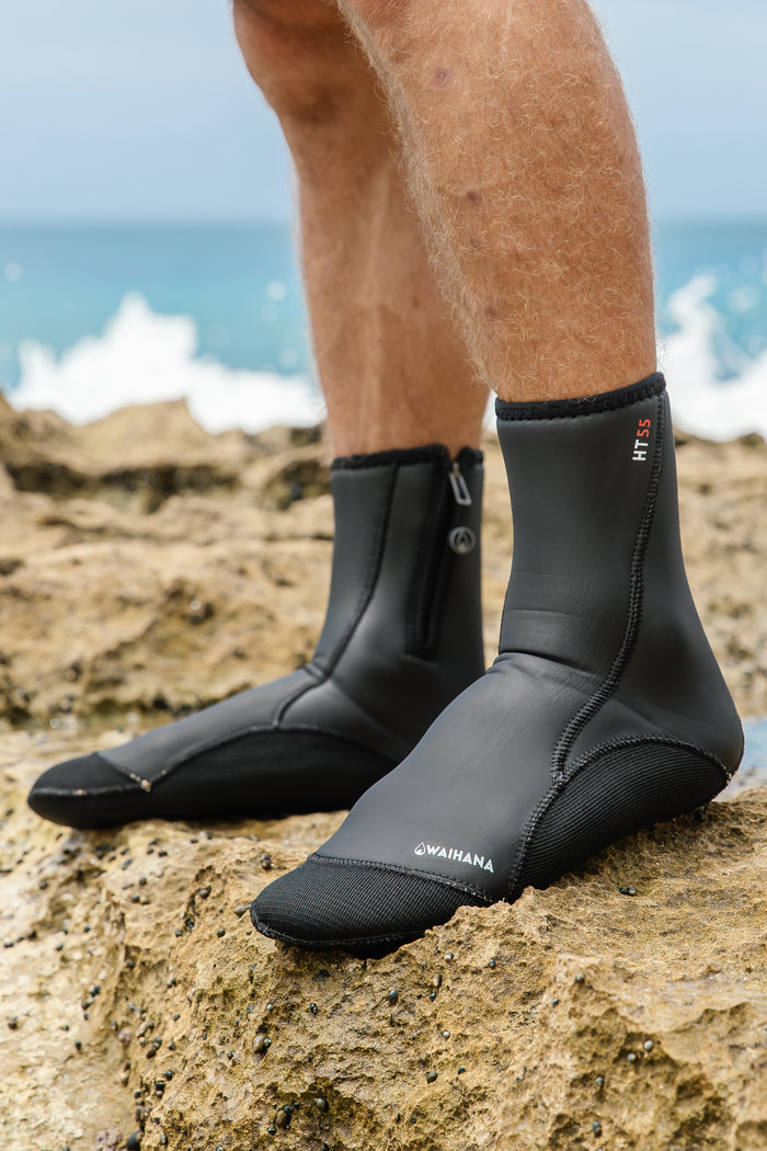 Close-up of a man's legs wearing black Waihana HT55 neoprene dive boots with zip closure, standing on coastal rocks with ocean waves in the background.