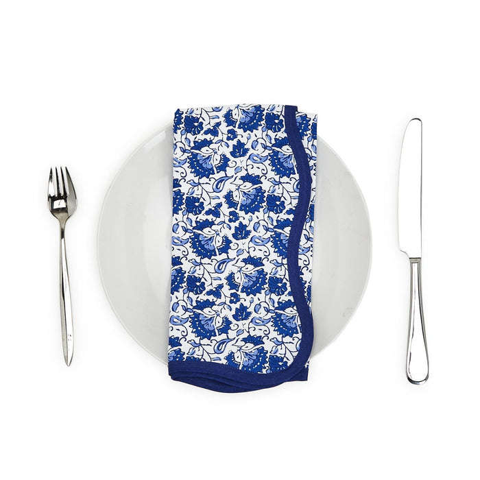 Blue floral cotton napkin with a scalloped blue trim, folded on a white plate with a fork and knife.