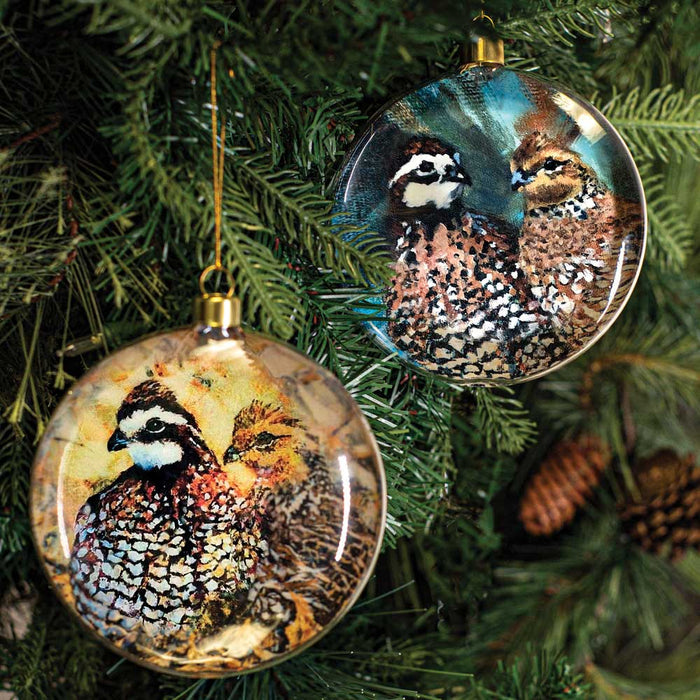 Two round glass ornaments featuring Sue Key quail artwork, hanging on a Christmas tree with gold accents.
