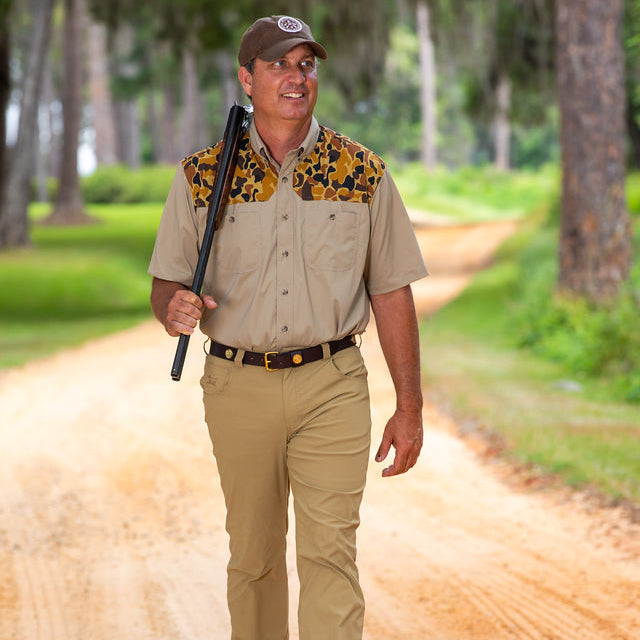 Man wearing a tan short-sleeve shooting shirt with camo shoulder patches and a matching cap while walking on a dirt path.