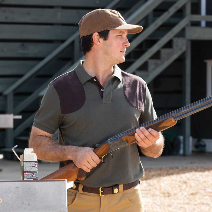 Man wearing an olive green Schoffel Cordoba polo shirt with brown suede shoulder patches and branded buttons while holding a shotgun.