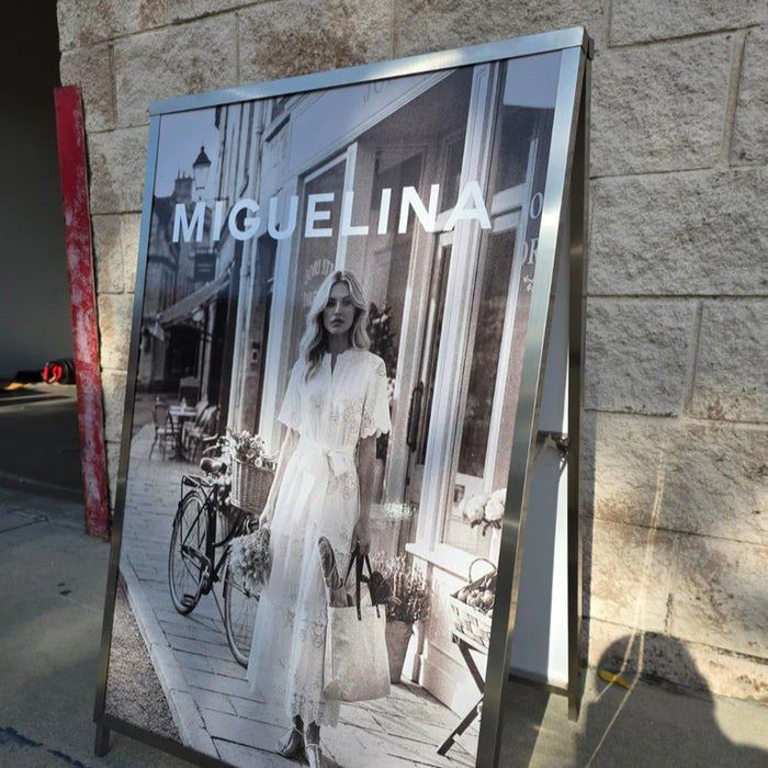 Angled view of an A-frame sign holder displaying a Miguelina fashion poster.
