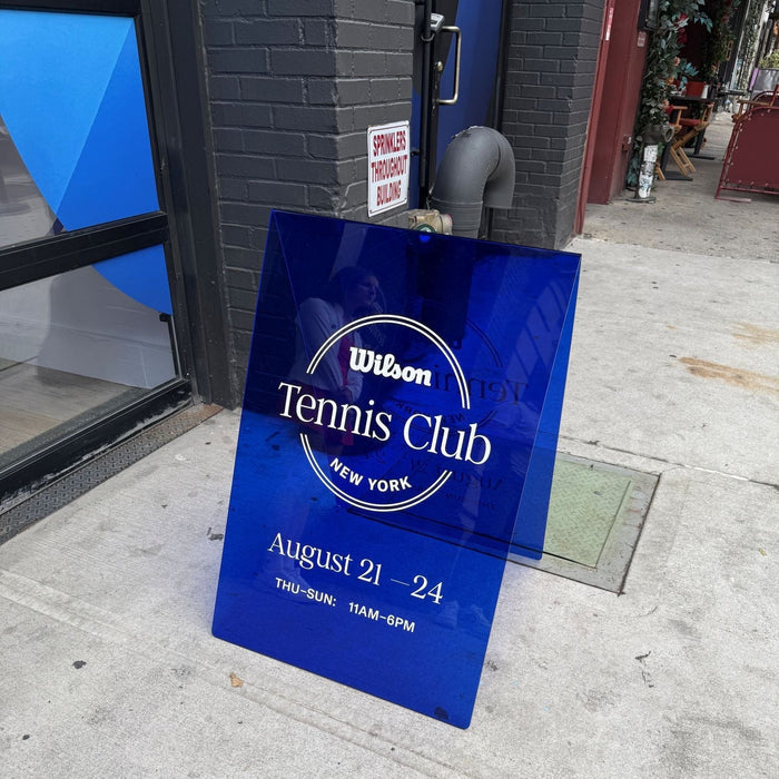 Transparent blue acrylic A-frame sign with Wilson Tennis Club lettering outside a storefront.