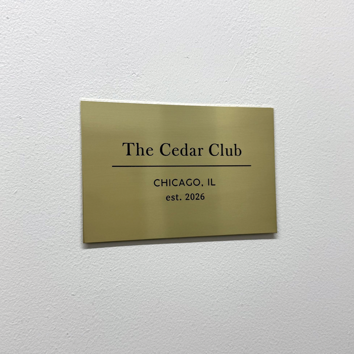 Brass gallery plaque with The Cedar Club lettering on a white wall.