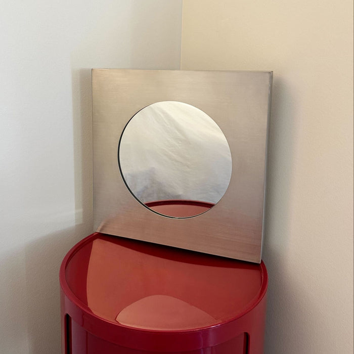 Square stainless steel Forma mirror standing on a red round table.