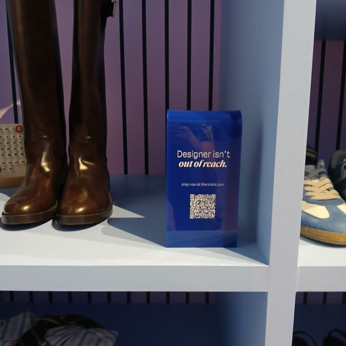 Blue acrylic sign with Designer Isn't Out of Reach lettering and a QR code beside boots.