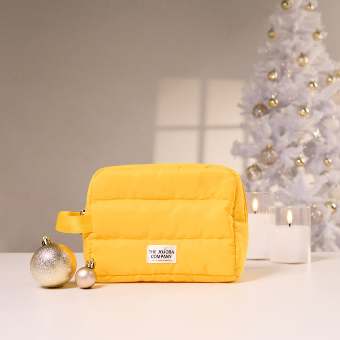 A bright yellow puffer-style cosmetic pouch with a small white brand label reading 'THE JOJOBA COMPANY' sits on a white surface. The scene is styled with holiday decor: two ornaments, two lit candles, and a white Christmas tree in the background.