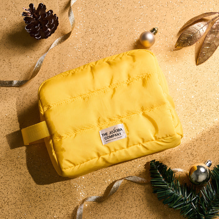 A bright yellow puffer-style cosmetic pouch with a small white brand label reading 'THE JOJOBA COMPANY,' styled on a glittery gold surface with holiday ornaments, ribbon, pine needles, and a pinecone.