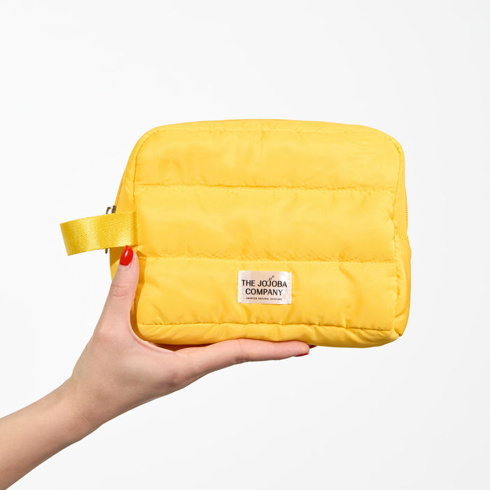 A hand with red nail polish holds a bright yellow puffer-style cosmetic pouch against a clean white background. The pouch has horizontal quilting, a matching yellow wrist strap, and a white brand label reading 'THE JOJOBA COMPANY'.