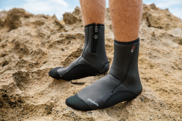 A man's legs wearing black Waihana HT55 neoprene dive socks standing on textured coastal rocks, showing both a zippered and zipperless version of the boot.