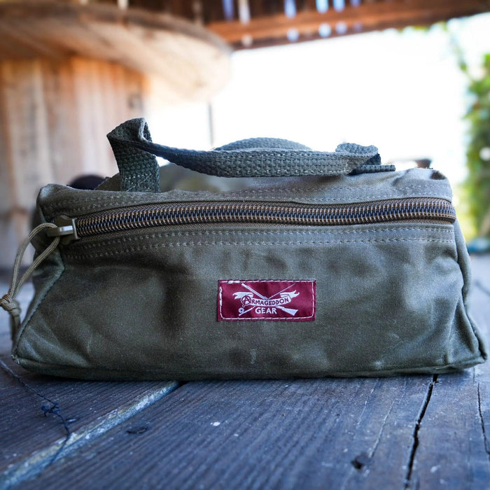 Olive green waxed canvas utility pouch with a top zipper, carrying handle, and Armageddon Gear logo patch.