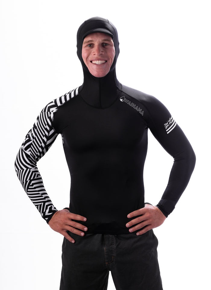 Male model wearing the Waihana Black Carvico Hooded Surf Rashguard with geometric black-and-white patterned sleeve and integrated hood, hands on hips against a white studio background.