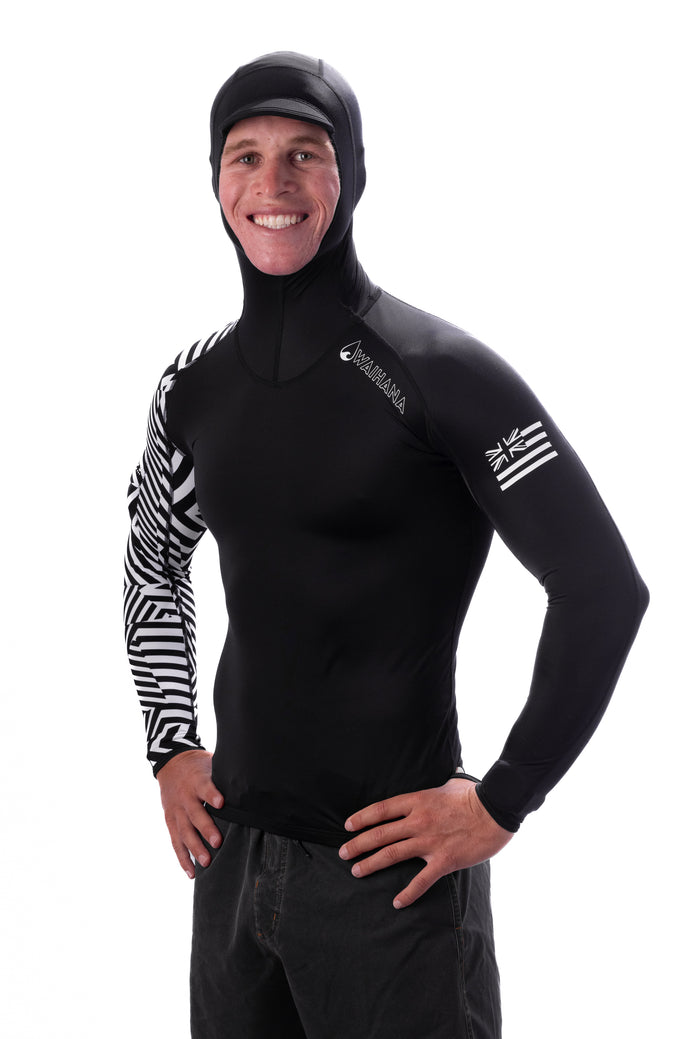 Smiling man wearing the Waihana Black Carvico Hooded Surf Rashguard featuring a black-and-white geometric print on the left sleeve and Hawaiian flag patch on the right arm, posed against a white background.