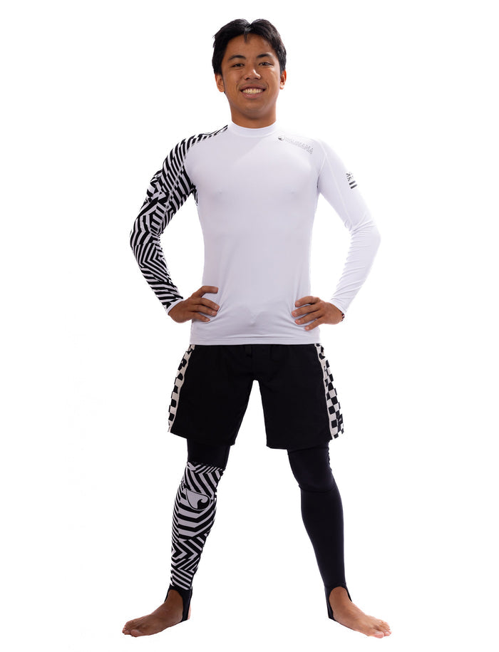 Male model wearing Waihana black Carvico Lycra leggings with bold geometric black-and-white print accents, paired with a white long-sleeve rashguard and black board shorts, standing confidently against a white background.