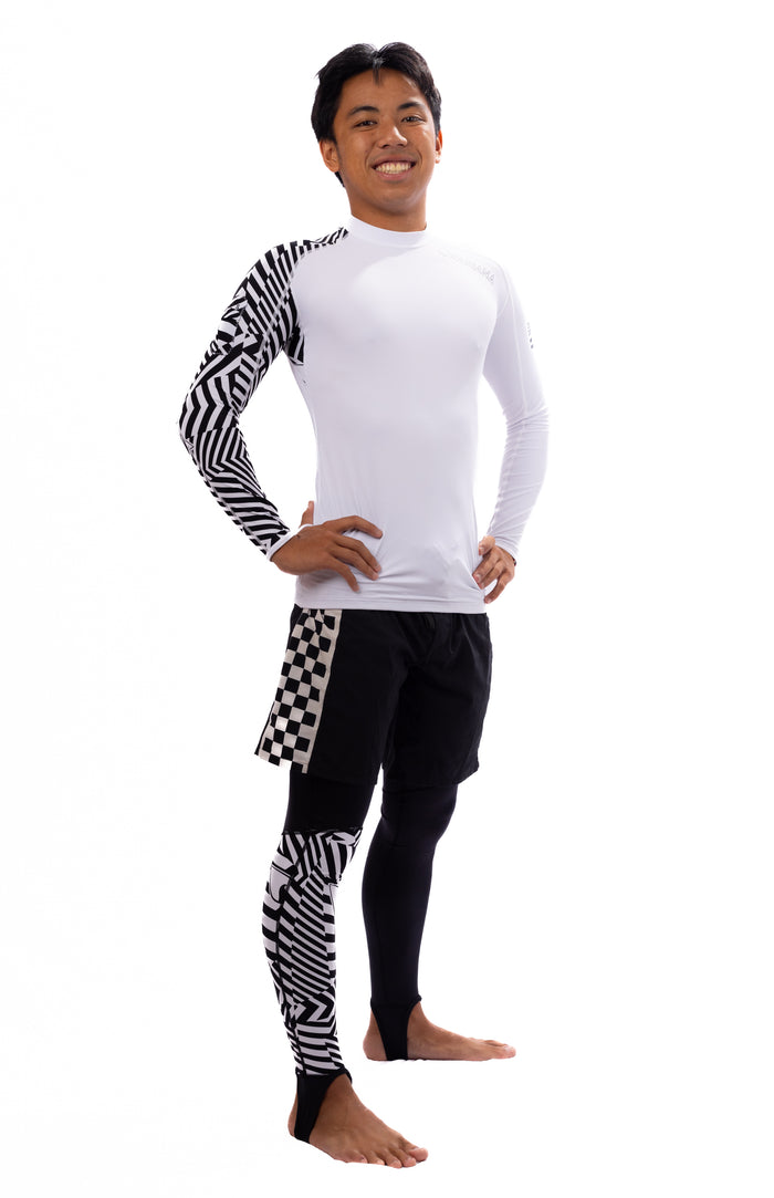 Male model smiling and wearing Waihana Black Carvico Lycra Leggings with geometric black-and-white pattern accents, paired with a white long-sleeve rashguard and black board shorts, on a white studio background.