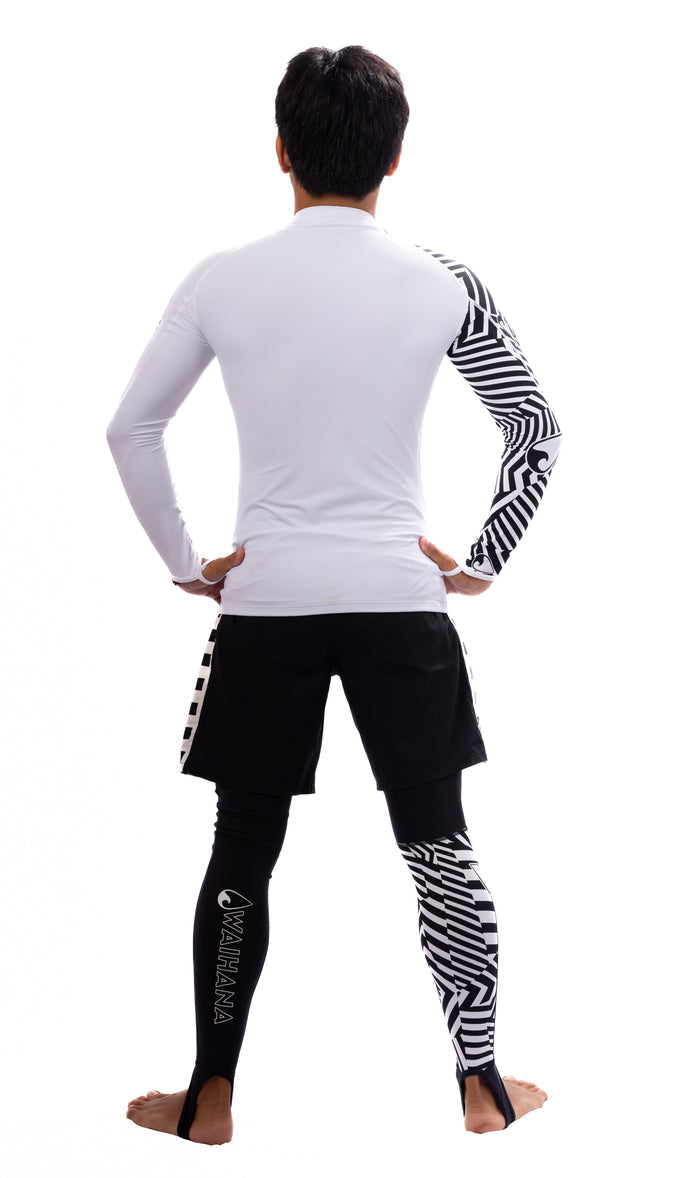 Back view of a male model wearing Waihana black compression leggings with bold geometric white stripe panels, paired with a white long-sleeve rashguard and black board shorts on a white background.