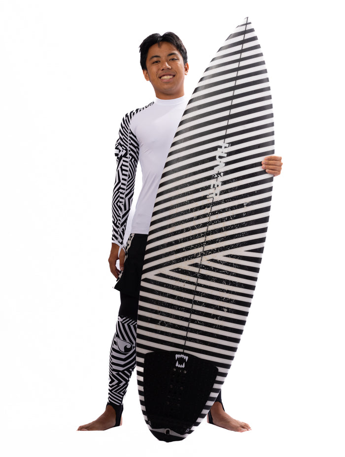 Smiling male model wearing Waihana's Black Carvico Lycra Leggings paired with a geometric-print rashguard, holding a black and white striped surfboard in a studio setting.