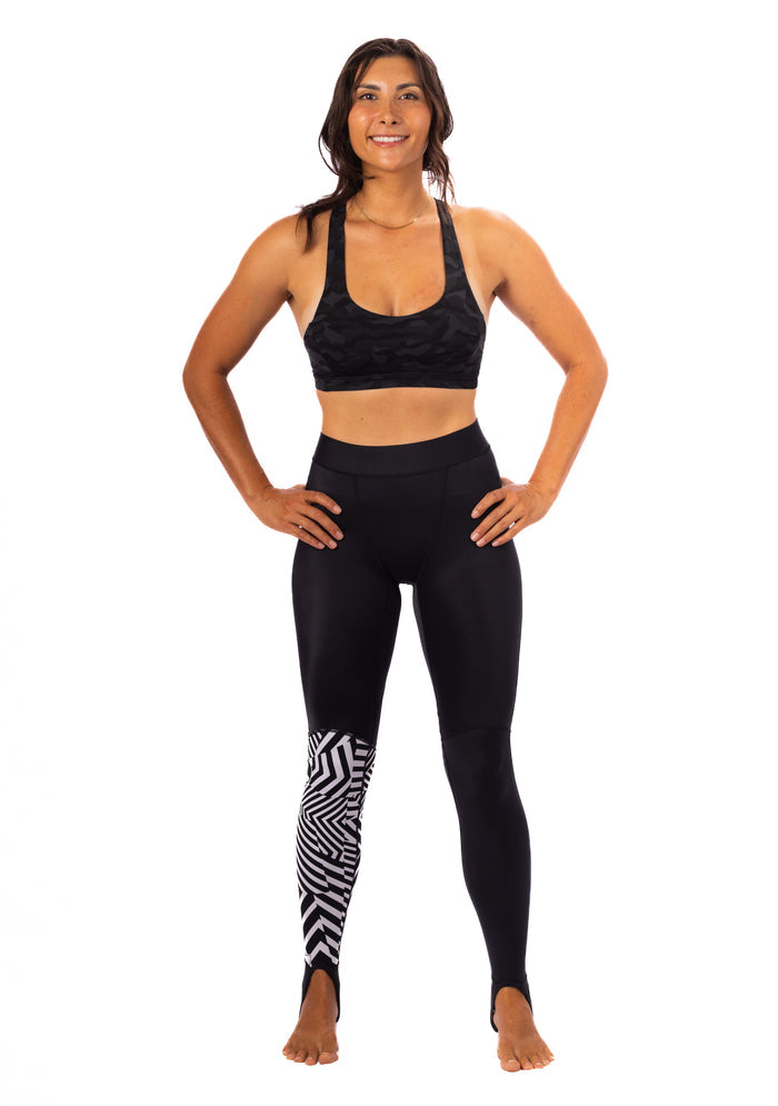 Woman wearing Waihana Black Carvico Lycra stirrup leggings with a geometric black-and-white chevron panel on the left leg, paired with a black camo sports bra, standing against a white background.