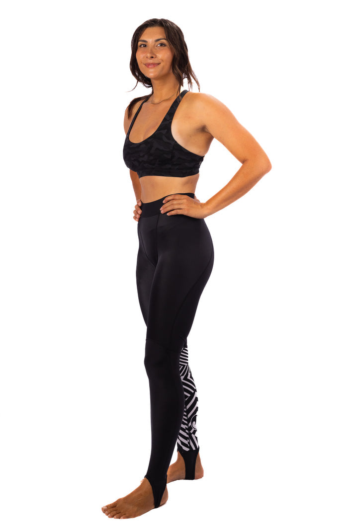 Woman modeling Waihana Black Carvico Lycra Leggings with white tribal graphic at the ankle, paired with a black sports bra, shown from a side angle against a white background.