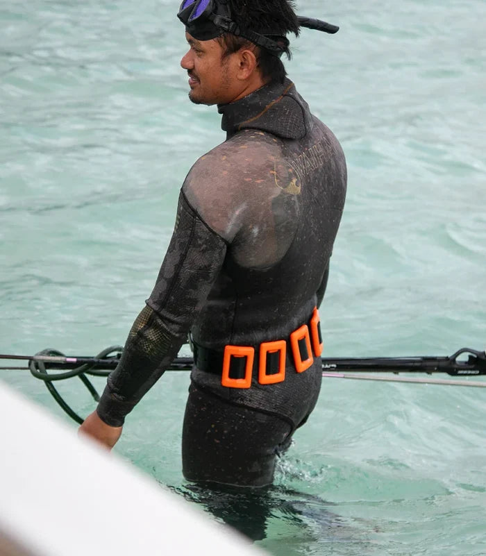 Male spearfisher standing waist-deep in clear turquoise ocean water wearing a Waihana camouflage wetsuit and an orange Clownfish dive weight belt, holding a speargun with mask pushed up on his cap.