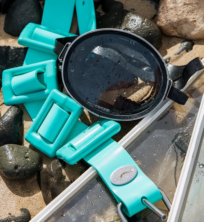 A teal Crystal River dive weight belt by Bright Buoy Diving with segmented plastic links and a metal fish-logo buckle resting on wet rocks and sand beside a Cressi smoked-lens dive mask.