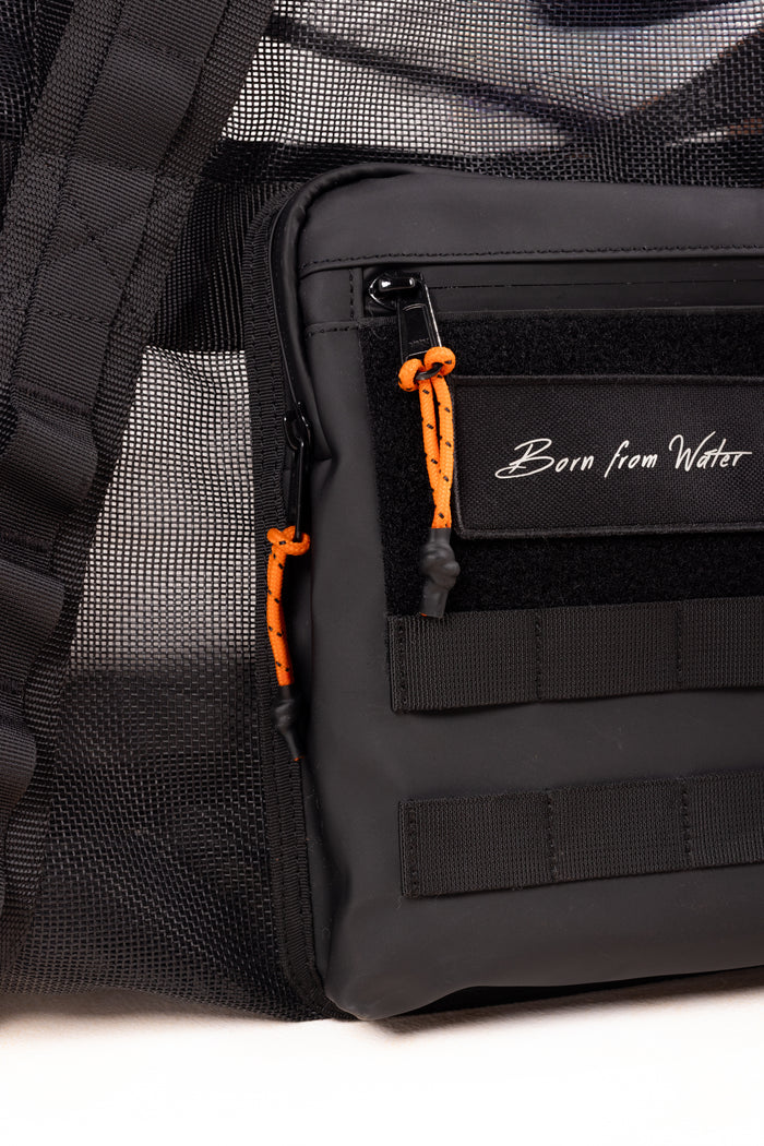 Close-up of a black Waihana gear bag featuring orange paracord zipper pulls, MOLLE webbing straps, mesh side panel, and a 'Born from Water' script logo on a velcro patch.