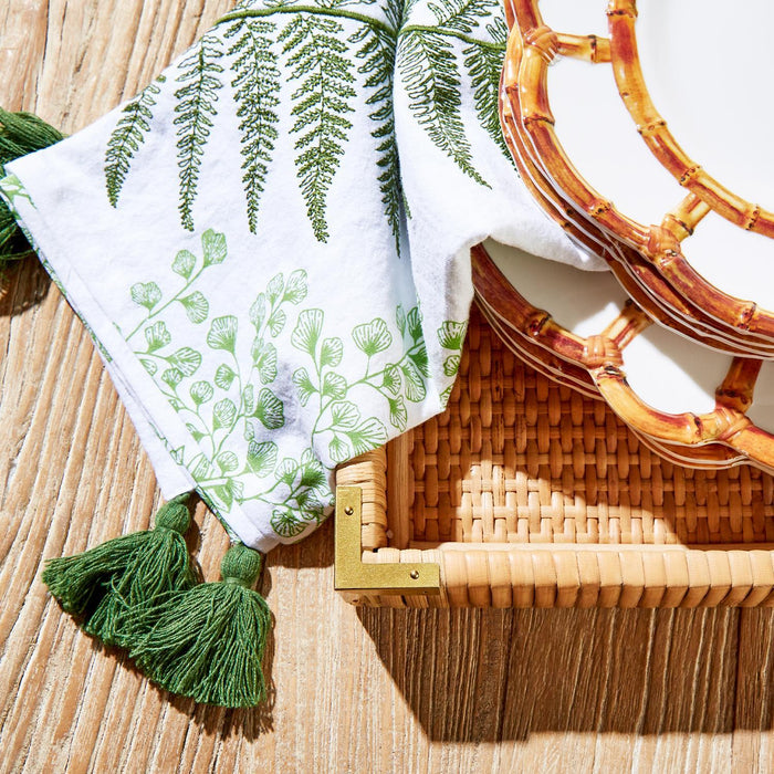 White cotton dish towel with green fern prints and tassel fringe, displayed on a woven rattan tray with bamboo-style plates.