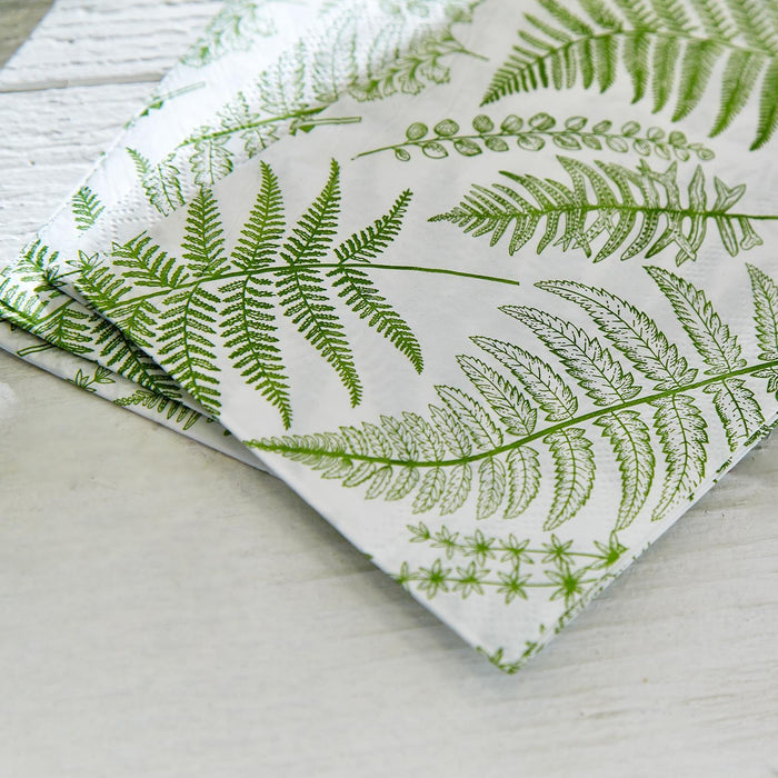 3-ply paper dinner napkin with a green fern botanical print on a white background, resting on a rustic white wood surface.