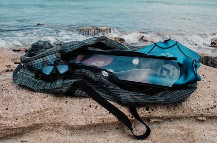 Gili Gear Freedive XL bag open on a coastal rock ledge, displaying a black freediving mask, long blade fins, and a teal wetsuit top, with ocean waves crashing in the background.