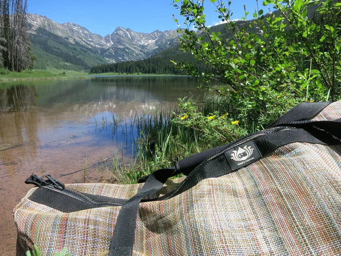 Gili Gear Freedive XL gear bag in multicolor woven fabric resting beside a serene alpine lake with pine forests and snow-capped mountains reflected in the water.