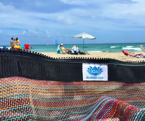 Close-up of the Gili Gear Freedive XL mesh bag in multicolor with a branded Gili Bags label on the zipper edge, with a sunny beach, ocean, and beachgoers visible in the background.