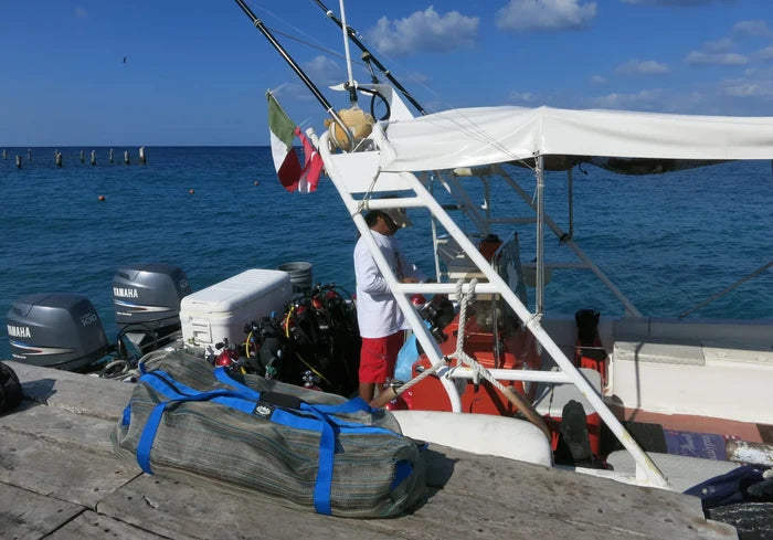 A Gili Gear Freedive XL mesh gear bag with blue straps rests on a dock beside a dive boat loaded with scuba equipment, with calm tropical ocean water in the background.