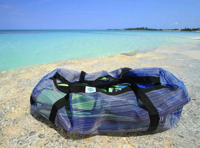 Gili Gear Freedive XL multi-color mesh gear bag packed with fins and freediving equipment resting on a rocky tropical shoreline with clear turquoise water behind it.
