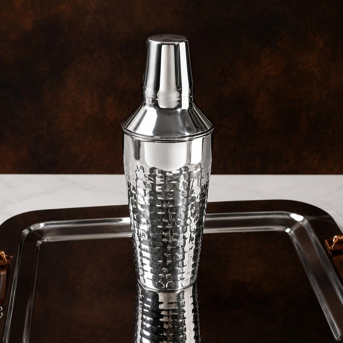 Silver hammered stainless steel cocktail shaker on a reflective metal tray.