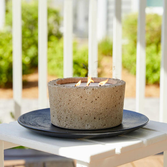 Large 32 oz outdoor candle in a textured beige hypertufa pot with three burning wicks, set on a black plate on a patio.