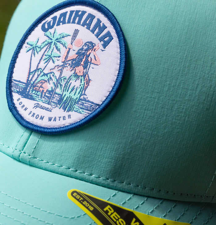 Close-up of a teal Waihana Hula Diver Hydro Hat with a circular embroidered patch depicting a Hawaiian figure holding a spear among palm trees and the phrase 'Born From Water, Hawaii'.