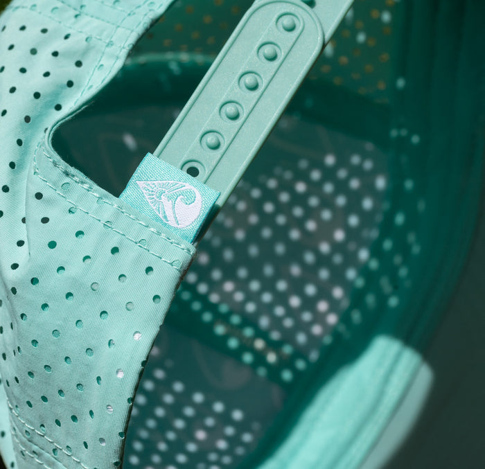 Close-up of the teal Waihana Hula Diver Hydro Hat showing perforated fabric, embroidered wave logo tag, and adjustable plastic back strap in matching teal.
