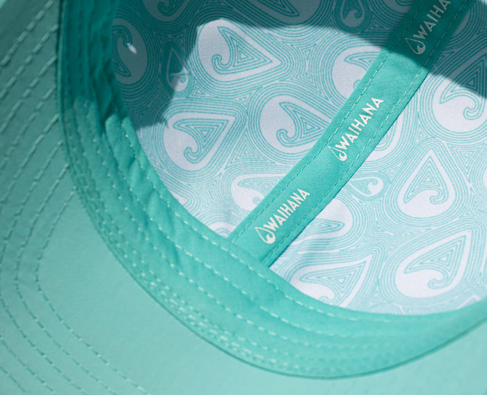 Close-up of the interior of a teal Waihana Hula Diver Hydro Hat, revealing a decorative branded lining with Waihana hook-and-wave motifs and a teal sweatband with repeated Waihana logo text.