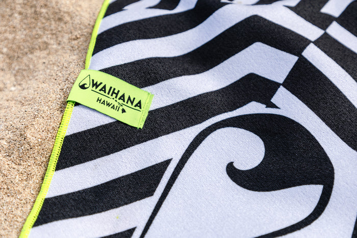 Close-up of the Waihana HydroTech Towel in black and white tribal wave print, showing the neon yellow Waihana Hawaii woven label and stitched edge resting on sand.