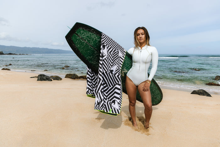 Woman in a white long-sleeve wetsuit stands barefoot on a sandy Hawaiian beach holding a dark green surfboard draped with the Waihana HydroTech Towel in black-and-white geometric print, ocean and mountains visible behind her.