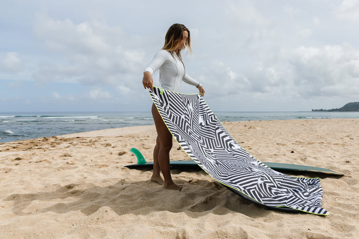 Woman in a white rashguard holding a large black-and-white geometric Waihana HydroTech Towel on a sandy beach with a surfboard and ocean in the background.