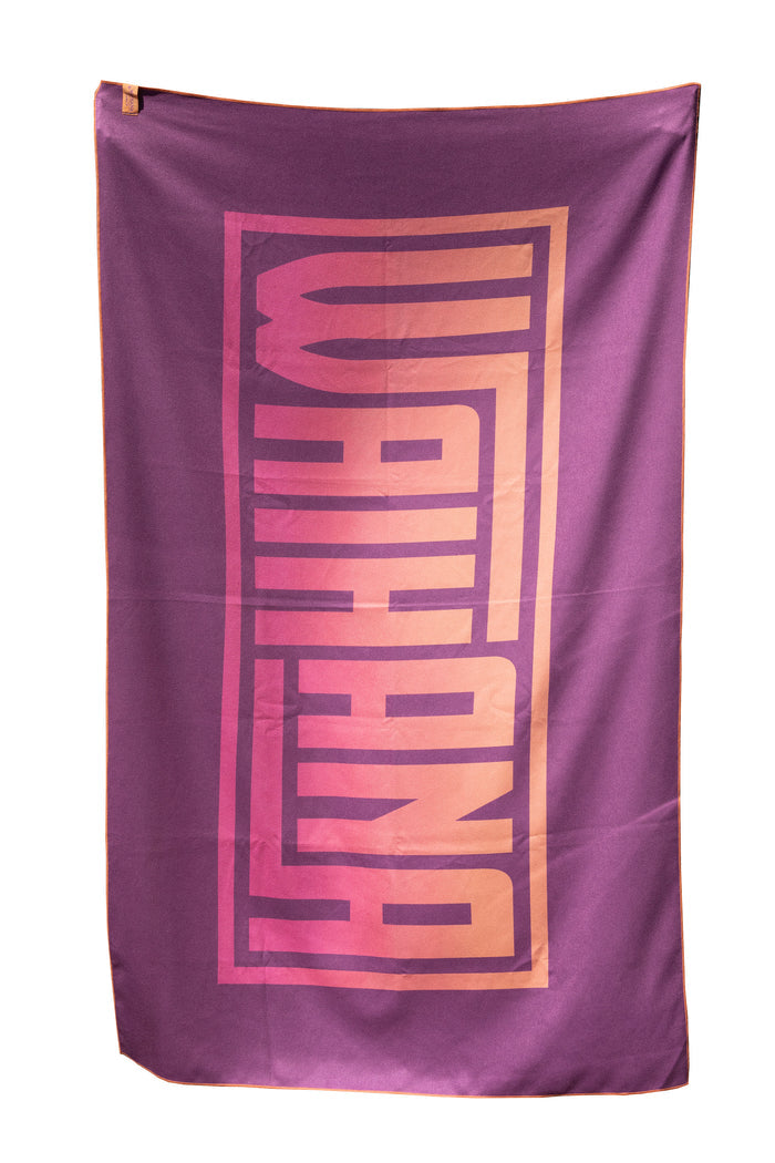 Waihana HydroTech Tropicam Towel in purple with bold pink-to-orange gradient brand lettering, shown laid flat on a white background.
