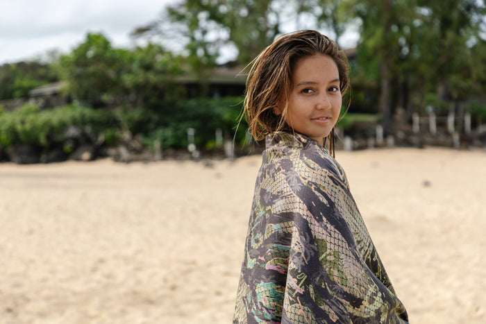 Young girl with wet hair wrapped in a Waihana HydroTech Tropicam patterned towel, smiling over her shoulder on a sandy beach with lush green trees in the background.