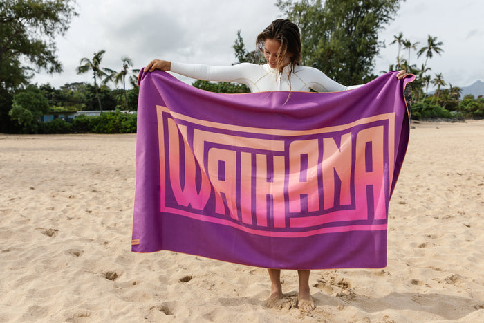 Woman in a white wetsuit holding up a large purple Waihana HydroTech Tropicam Towel with bold pink and peach logo text on a sandy beach with palm trees in the background.