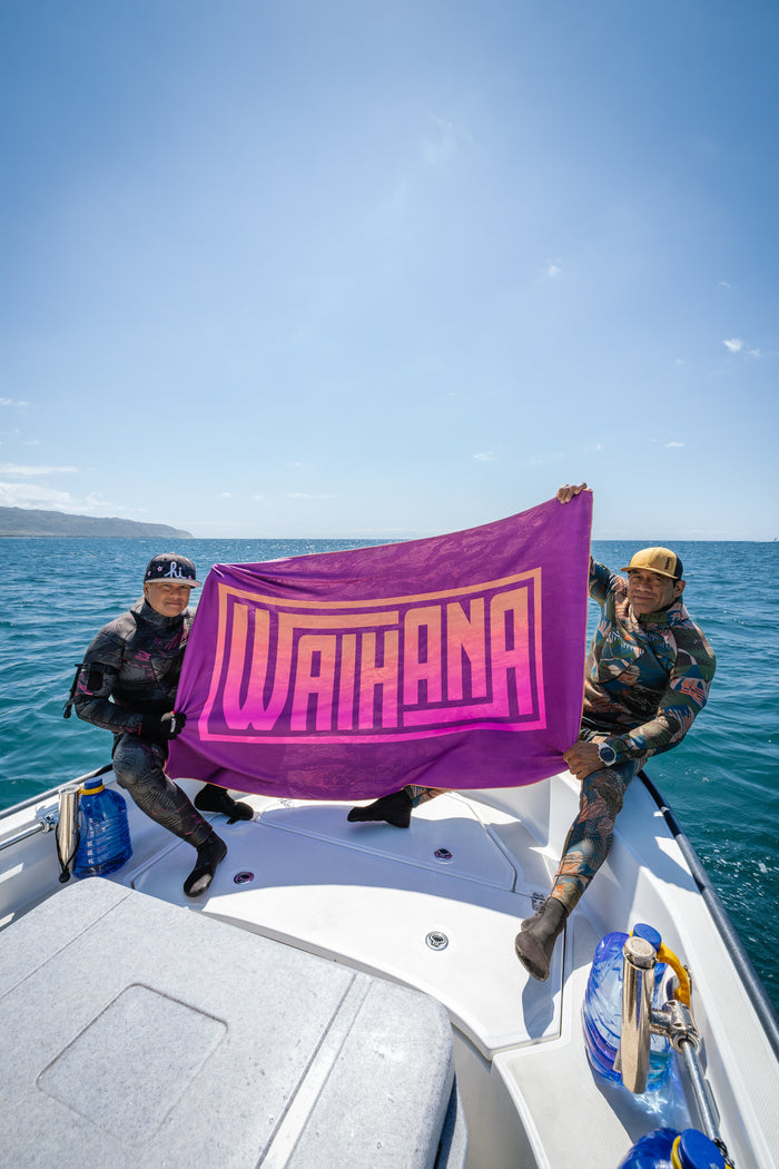 Two spearfishers in wetsuits hold up a large purple Waihana HydroTech Tropicam Towel on the bow of a boat with blue ocean and coastline in the background.