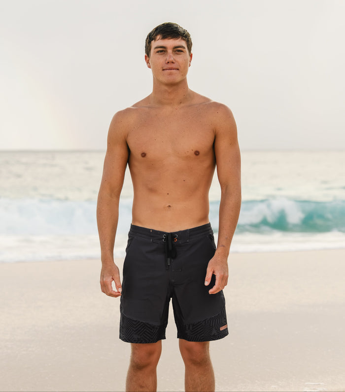 Shirtless male model wearing Waihana Jet Black W1-PRO board shorts standing on a sandy beach with ocean waves breaking in the background.