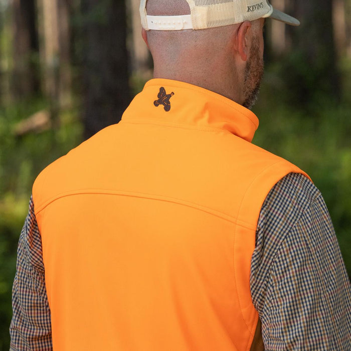 Bright orange windproof softshell vest with a high collar and logo, worn over a plaid shirt in a wooded outdoor setting.