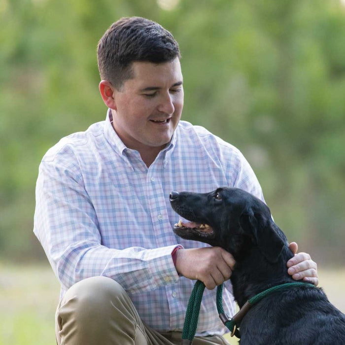 Man wearing a wrinkle-free pink and blue plaid button-down cotton shirt while petting a black dog outdoors.