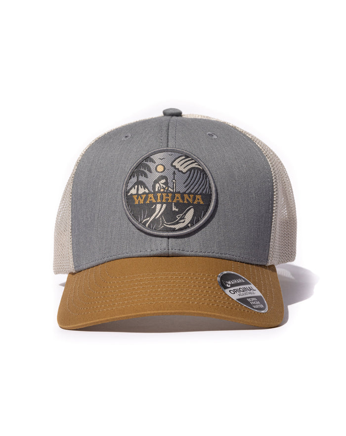 Waihana Lagoon Diver Trucker hat in grey with tan mesh back and caramel brim, featuring a circular woven patch depicting a spearfisher underwater with a whale, ocean waves, palm trees, and gold Waihana lettering.