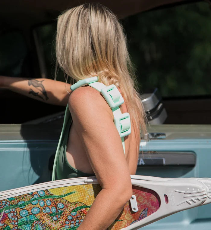 Blonde woman seen from behind wearing a mint green buckle-strap bikini top, holding vibrant printed freediving fins beside a boat with an outboard motor.